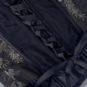 May include: Close-up of a black corset with intricate details. The corset features a lace-up back, delicate silver sequin and bead embellishments, and a sheer mesh overlay. The satin ribbon ties add a touch of elegance to this garment.