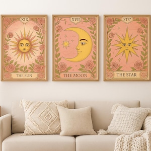 Op de afbeelding: Drie ingelijste tarotkaarten met de Zon, Maan en Ster. Elke kaart heeft een roze achtergrond met bloemen en botanische elementen. De Zon-kaart toont een zon met een gezicht, de Maan-kaart toont een halve maan en de Ster-kaart heeft een ster.