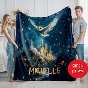May include: A navy blue blanket featuring a white owl in flight above a castle with golden lights. The blanket has the name "MICHELLE" in gold lettering. The blanket is held by two people.