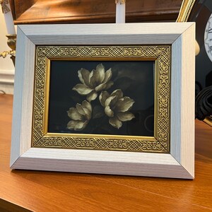 May include: A framed floral print showing three white flowers with yellow centres against a dark backdrop. The frame has a light grey outer border and a gold inner border with an ornate design. The artwork is displayed on a wooden surface.