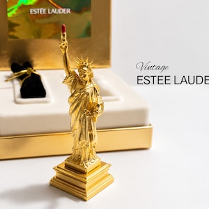 May include: A gold-toned Statue of Liberty figurine holding a red lipstick in its raised hand, presented in a gold box with white velvet lining. The text "Vintage ESTEE LAUDER" is printed on the right, and "ESTEE LAUDER" appears on the box lid.