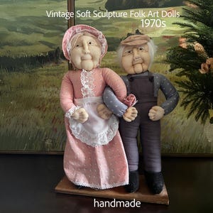 May include: Vintage soft sculpture folk art dolls from the 1970s. The handmade dolls depict an elderly couple. The woman wears a pink dress and apron, while the man wears overalls and a cap. The dolls are standing on a wooden base.