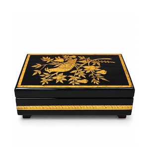 May include: A rectangular black box with a hinged lid featuring a gold-colored illustration of a bird perched on a branch with leaves and fruit. The lid has a gold border, and the front side displays a decorative gold scroll pattern.