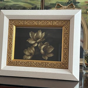 May include: A framed floral print showing three cream-coloured flowers on a dark backdrop. The frame has a white outer edge and a gold inner border with an intricate design. The artwork is displayed on a table.