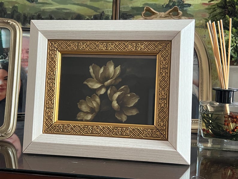 May include: A framed floral print showing three cream-coloured flowers on a dark backdrop. The frame has a white outer edge and a gold inner border with an intricate design. The artwork is displayed on a table.