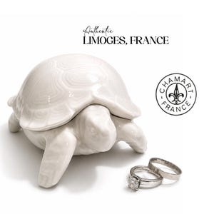 May include: A white porcelain turtle-shaped trinket box with a textured shell, accompanied by two silver-toned rings. The text reads Authentic LIMOGES, FRANCE and CHAMART FRANCE with a fleur-de-lis symbol. The object is glossy and smooth.