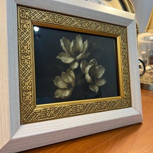 May include: A framed floral art print showing three white and beige flowers on a dark background. The frame has a gold ornate inner border and a light wood outer frame. The artwork is displayed on a wooden surface.