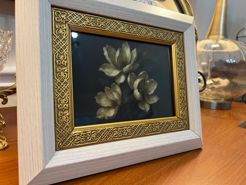 May include: A framed floral art print showing three white and beige flowers on a dark background. The frame has a gold ornate inner border and a light wood outer frame. The artwork is displayed on a wooden surface.