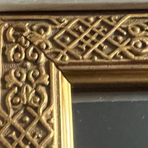 May include: Detail of a gold-coloured picture frame corner with intricate, repeating patterns. The frame has a light-coloured outer edge. The design features scrolling and geometric shapes, typical of a decorative frame.