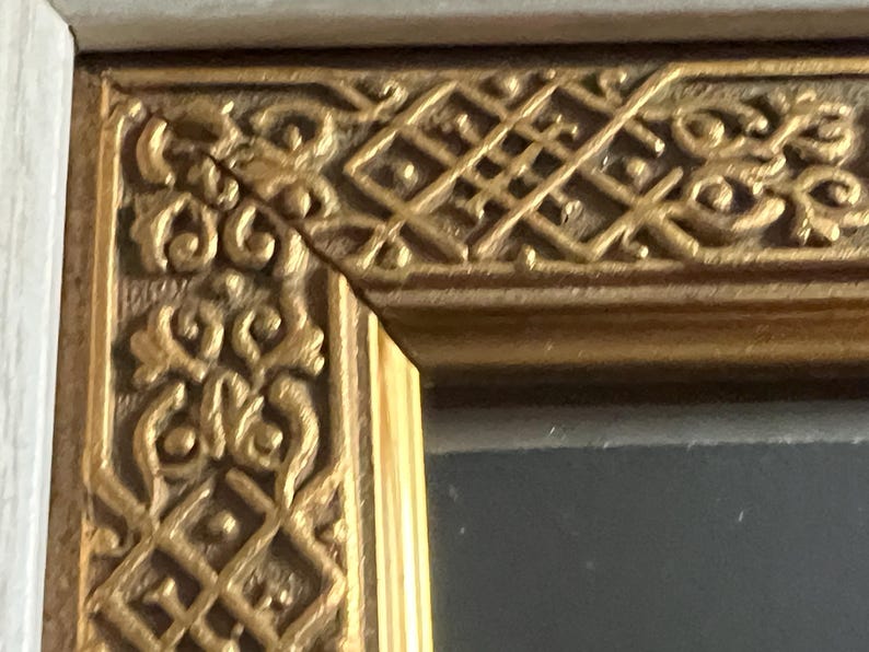 May include: Detail of a gold-coloured picture frame corner with intricate, repeating patterns. The frame has a light-coloured outer edge. The design features scrolling and geometric shapes, typical of a decorative frame.