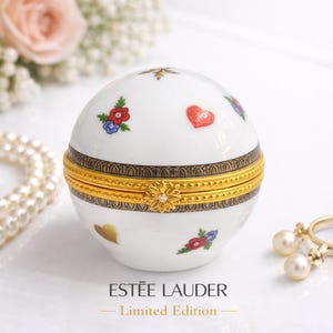 May include: A white, spherical trinket box with a gold-colored band and floral and heart designs. The box has the text "ESTÉE LAUDER - Limited Edition". The box is decorated with red, blue, and gold accents.