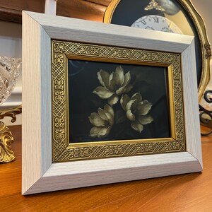 May include: A framed floral print with three cream-coloured flowers on a dark background. The frame has a light wood finish and a gold ornate inner border. The artwork is displayed on a wooden surface.