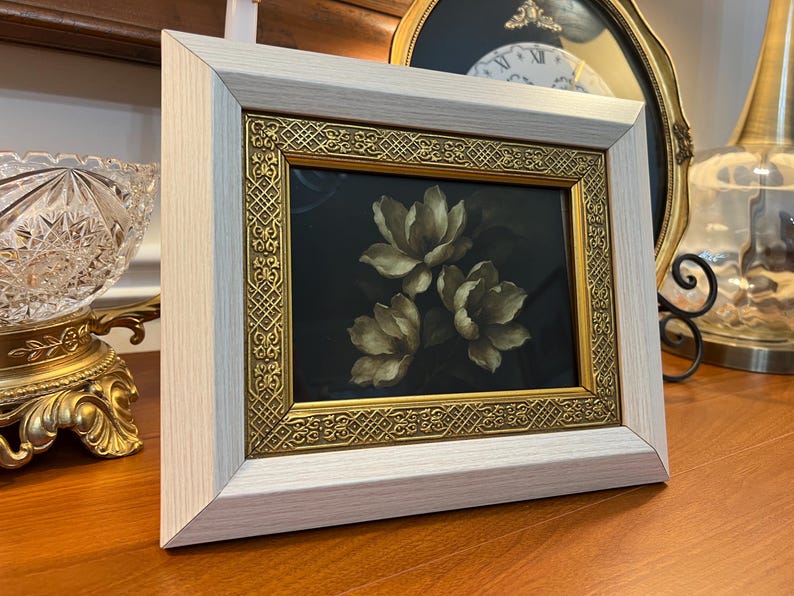 May include: A framed floral print with three cream-coloured flowers on a dark background. The frame has a light wood finish and a gold ornate inner border. The artwork is displayed on a wooden surface.