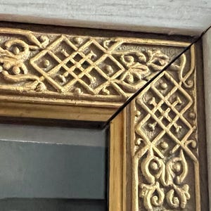 May include: Close-up of a decorative picture frame corner. The frame features ornate gold-coloured relief carvings with geometric and floral patterns. The frame's outer edge is a light, neutral colour.