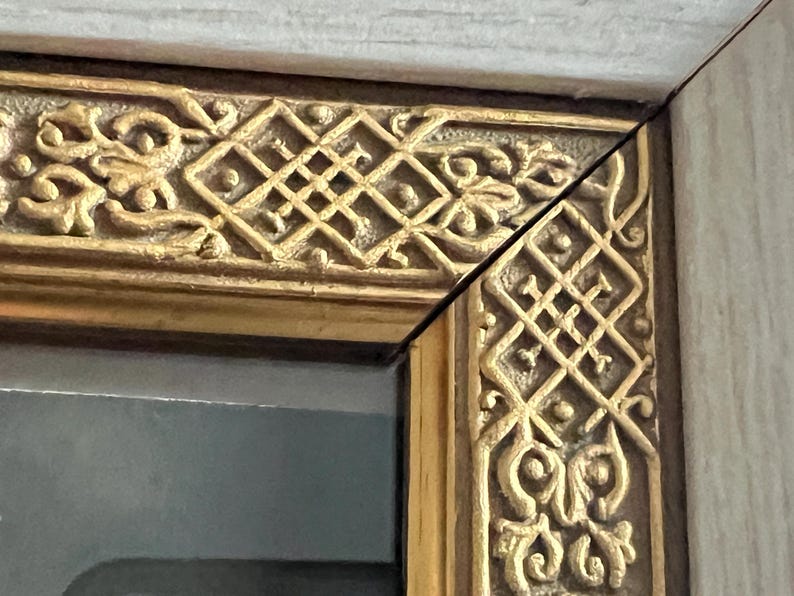 May include: Close-up of a decorative picture frame corner. The frame features ornate gold-coloured relief carvings with geometric and floral patterns. The frame's outer edge is a light, neutral colour.