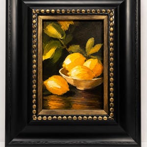 Puede incluir: Una pintura al &oacute;leo de limones de color amarillo brillante en un cuenco blanco sobre un fondo oscuro. La obra est&aacute; enmarcada en un marco negro grueso con un borde interior dorado que presenta una fila decorativa de peque&ntilde;os tachones met&aacute;licos.