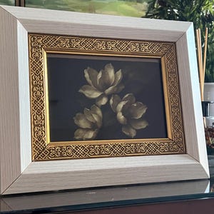 May include: A framed floral print showing three white flowers on a dark backdrop. The frame has a light wood grain finish with a gold filigree border. The artwork is displayed on a reflective surface.
