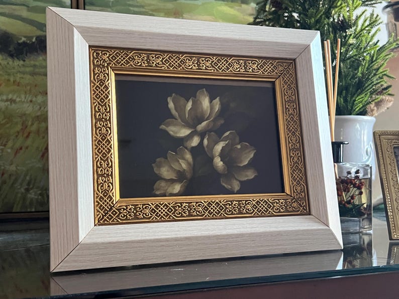 May include: A framed floral print showing three white flowers on a dark backdrop. The frame has a light wood grain finish with a gold filigree border. The artwork is displayed on a reflective surface.