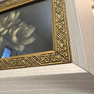 May include: Close-up of a framed artwork featuring a floral design. The frame has a gold-coloured, ornate border and a light wood-grain finish. The artwork inside the frame depicts large, cream-coloured flowers.