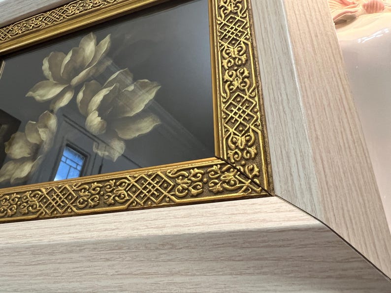 May include: Close-up of a framed artwork featuring a floral design. The frame has a gold-coloured, ornate border and a light wood-grain finish. The artwork inside the frame depicts large, cream-coloured flowers.