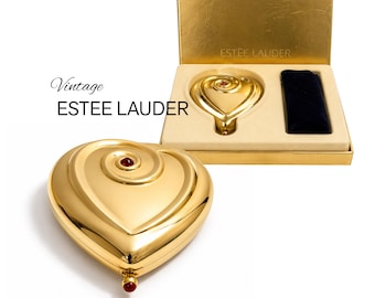 May include: A gold-toned heart-shaped Estée Lauder compact with a spiral design and a small red stone accent. The set includes a matching gold compact inside a rectangular gold box with a cream-colored insert and a dark blue velvet pouch.