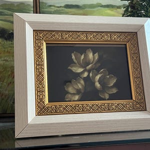 May include: A framed floral print showing three cream-coloured flowers against a dark background. The frame has a light wood-grain finish with a gold decorative inner border. The artwork is displayed on a reflective surface.