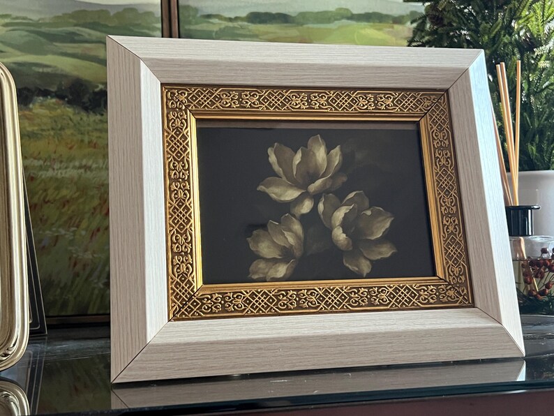 May include: A framed floral print showing three cream-coloured flowers against a dark background. The frame has a light wood-grain finish with a gold decorative inner border. The artwork is displayed on a reflective surface.