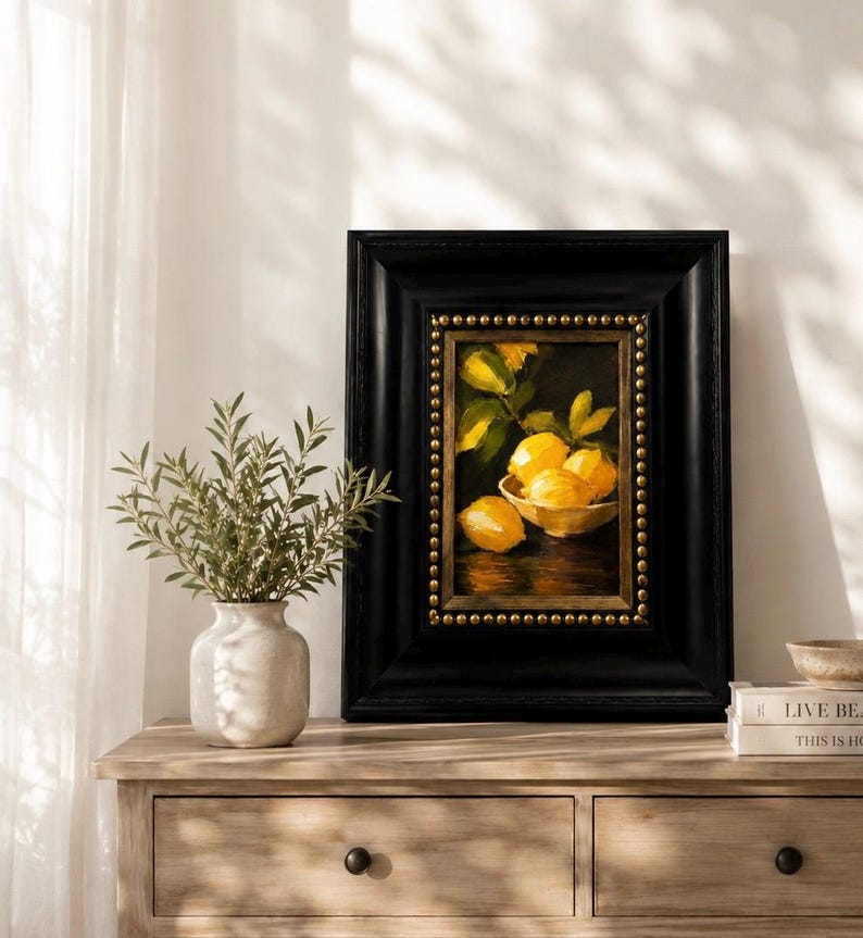 Puede incluir: Un &oacute;leo enmarcado que representa un cuenco de limones amarillos sobre una superficie oscura, dentro de un marco negro ancho con adornos de tachuelas doradas. La obra se exhibe sobre una c&oacute;moda de madera junto a un jarr&oacute;n de cer&aacute;mica blanca.