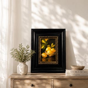 Puede incluir: Un &oacute;leo enmarcado que representa un cuenco de limones de color amarillo brillante sobre un fondo oscuro. La obra est&aacute; dentro de un marco negro grueso con adornos de tachuelas doradas. La pintura captura los c&iacute;tricos en un cuenco.