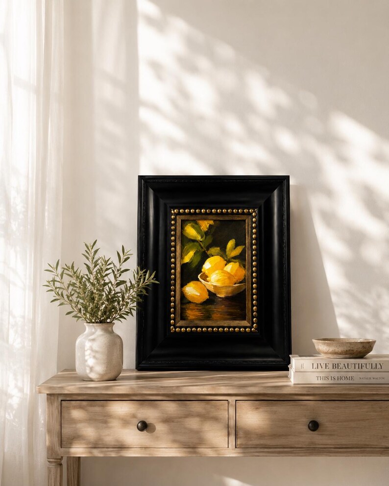 Puede incluir: Un &oacute;leo enmarcado que representa un cuenco de limones de color amarillo brillante sobre un fondo oscuro. La obra est&aacute; dentro de un marco negro grueso con adornos de tachuelas doradas. La pintura captura los c&iacute;tricos en un cuenco.