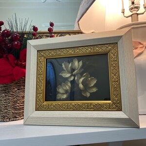 May include: A framed floral print with a light wood frame and gold trim. The artwork features three white flowers against a dark background. A wicker basket with red berries and a white lamp with a pink bow are also visible.