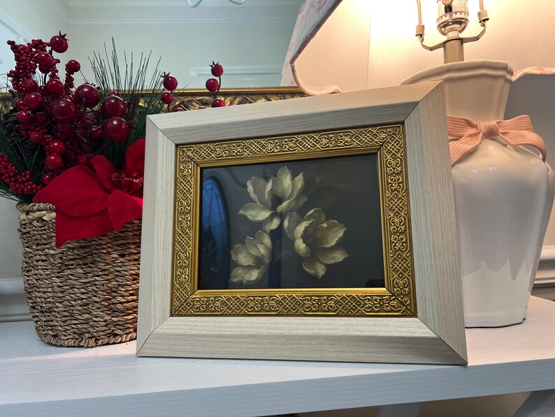 May include: A framed floral print with a light wood frame and gold trim. The artwork features three white flowers against a dark background. A wicker basket with red berries and a white lamp with a pink bow are also visible.