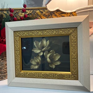 May include: A framed floral print showing three cream-coloured flowers against a dark background. The frame has a light wood finish with a gold ornate inner border. The artwork is displayed on a white surface.
