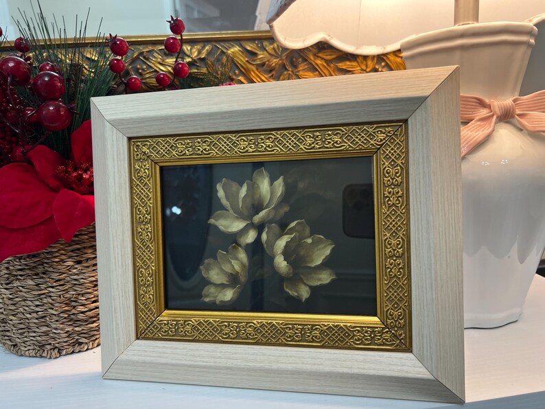 May include: A framed floral print showing three cream-coloured flowers against a dark background. The frame has a light wood finish with a gold ornate inner border. The artwork is displayed on a white surface.