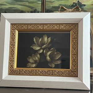 May include: A framed floral art print showing three cream-coloured flowers against a dark background. The frame has a white outer border and an ornate gold inner border. The artwork is displayed on a dark surface.