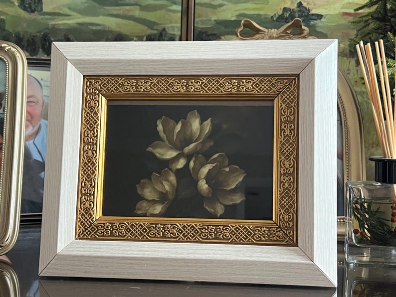 May include: A framed floral art print showing three cream-coloured flowers against a dark background. The frame has a white outer border and an ornate gold inner border. The artwork is displayed on a dark surface.