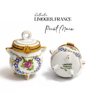 May include: Two white Limoges porcelain trinket boxes with gold detailing. One is upright, the other on its side. Both have hand-painted floral patterns with pink roses and blue accents. The text "Authentic LIMOGES, FRANCE Peint Main" is visible.