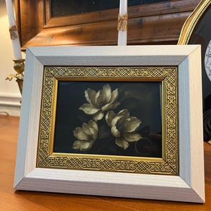 May include: A framed floral print featuring three white and gold flowers against a dark background. The frame has a light wood finish with a gold filigree border. The artwork is displayed on a wooden surface.