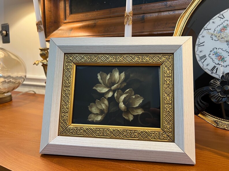May include: A framed floral print featuring three white and gold flowers against a dark background. The frame has a light wood finish with a gold filigree border. The artwork is displayed on a wooden surface.