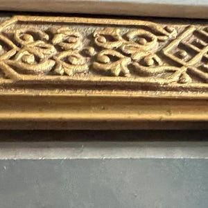 May include: Detail of a decorative gold frame with ornate patterns. The frame displays repeating floral and geometric designs, with a textured surface and a darker, aged finish. The frame is likely part of a larger artwork.