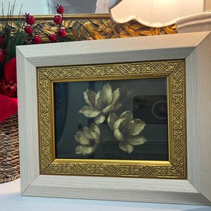 May include: A framed floral print showing three cream-coloured flowers against a dark background. The frame has a light wood finish with a gold ornate inner border. The artwork is displayed on a white surface.
