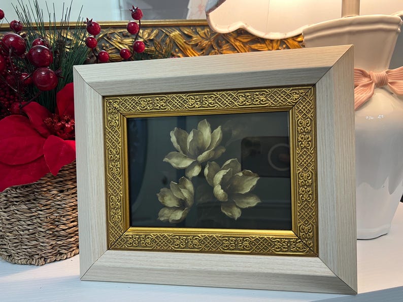 May include: A framed floral print showing three cream-coloured flowers against a dark background. The frame has a light wood finish with a gold ornate inner border. The artwork is displayed on a white surface.