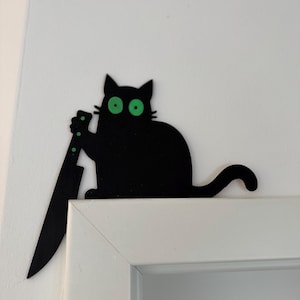 Könnte beinhalten: Schwarze Katzen-Dekoration mit grünen Augen und einem großen Messer. Die Katze sitzt auf einem weißen Türrahmen und erzeugt eine humorvolle und leicht bedrohliche Ästhetik. Das Messer hat grüne Punkte.
