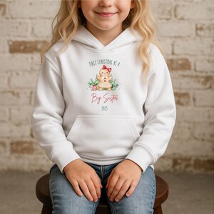 Könnte beinhalten: Weißer Kapuzenpullover mit Fronttasche. Der Pullover zeigt den Text "First Christmas as a Big Sister 2025" über einer Lebkuchenmännchen-Grafik mit roter Schleife. Das Design ist im Aquarellstil gehalten.