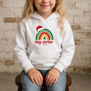Könnte beinhalten: Weißer Kapuzenpullover mit Regenbogen-Grafik, Weihnachtsmütze und dem Schriftzug "big sister 2026" in Rot. Das Design beinhaltet einen Weihnachtsbaum. Der Kapuzenpullover hat eine Vordertasche und wird von einem Kind getragen.