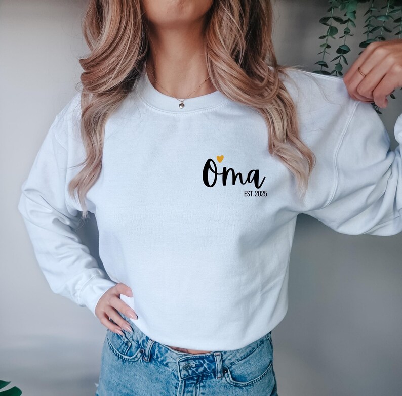 K&ouml;nnte beinhalten: Wei&szlig;es Langarm-Sweatshirt mit dem Wort "Oma" in schwarzer Schrift, einem kleinen gelben Herz und dem Text "EST. 2025". Das Sweatshirt hat einen l&auml;ssigen Stil.