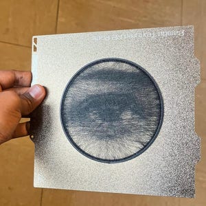 Könnte beinhalten: Eine quadratische, metallische 3D-Drucker-Bauplatte mit texturierter silberner Oberfläche und einem kreisförmigen schwarzen Filamentdruck in der Mitte. Die Platte trägt den Text Bambu Textured PEI Plate in der oberen rechten Ecke.