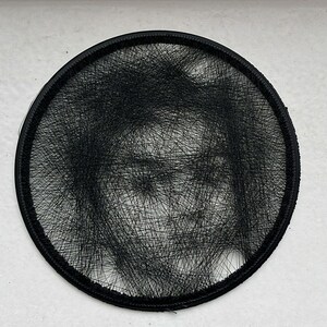 以下が含まれることがあります： 複雑な黒い糸のアートで作成された肖像画が入った円形の黒いフレーム。密集して重なり合う線が人物の顔の細部を形成し、丸い縁取りの中にモノクロの画像を作り出しています。