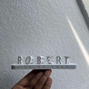 以下が含まれることがあります： 白い3Dプリント製のネームプレート。浮き彫りになった大文字の「ROBERT」という名前と、水平なベース部分に小さなフォントで刻印された「26.08.1997」という日付が特徴で、全体が単一の白い固体として構成されています。