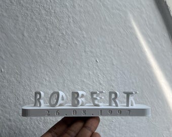 May include: A white 3D printed name plaque featuring the name ROBERT in raised capital letters, with the date 26.08.1997 embossed in a smaller font on the horizontal base platform, presented as a single solid white object.